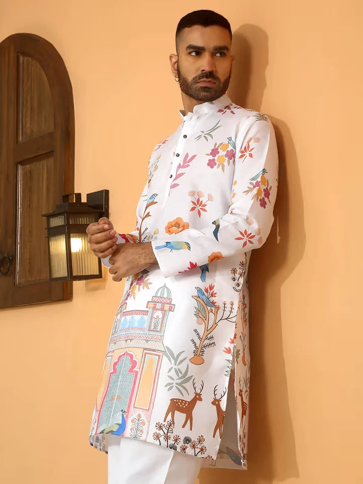 A man wearing a light peach kurta with intricate golden patterns and colorful landscape prints near the cuffs and hem, paired with white churidar pants and brown formal shoes, standing against a peach-colored background.