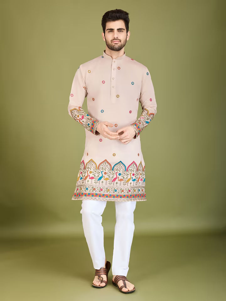 Man wearing a beige modal printed kurta with vibrant peacock and floral motifs, paired with white pants, standing beside a wooden stool.