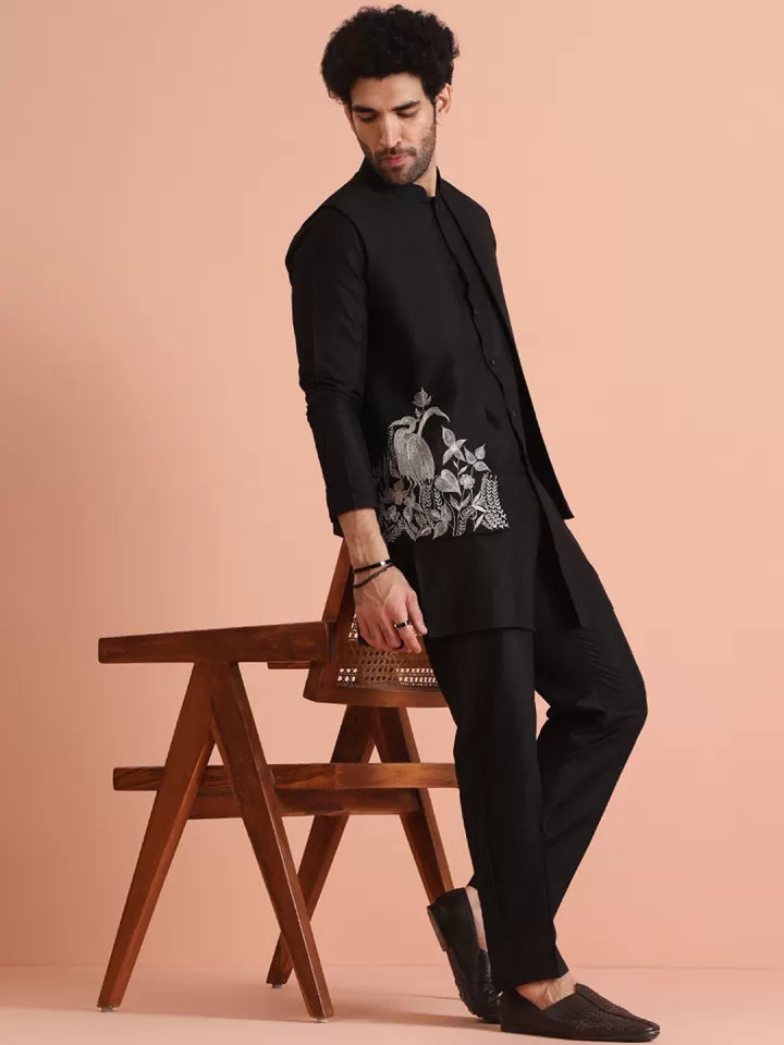 Man in a black outfit with a white design sitting on a wooden chair against a beige background