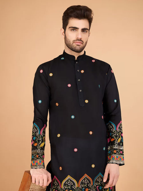 “Man wearing a black printed kurta with multicolor traditional patterns, paired with white pants, standing beside a wooden chair.”