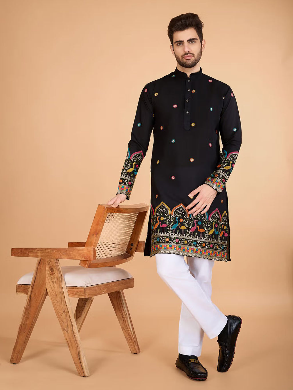 Man wearing a black embroidered kurta with white pants standing next to a wooden chair on a beige background