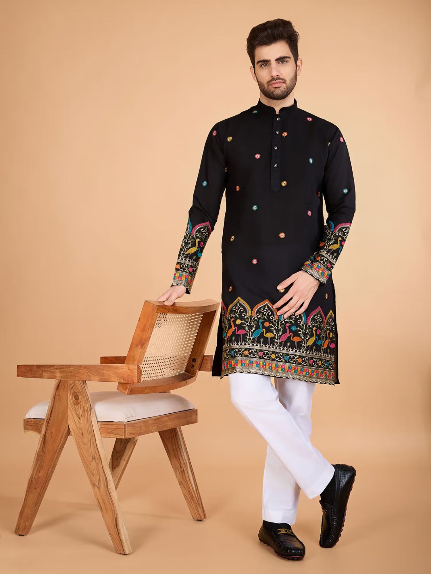 Man wearing a black embroidered kurta with white pants standing next to a wooden chair on a beige background