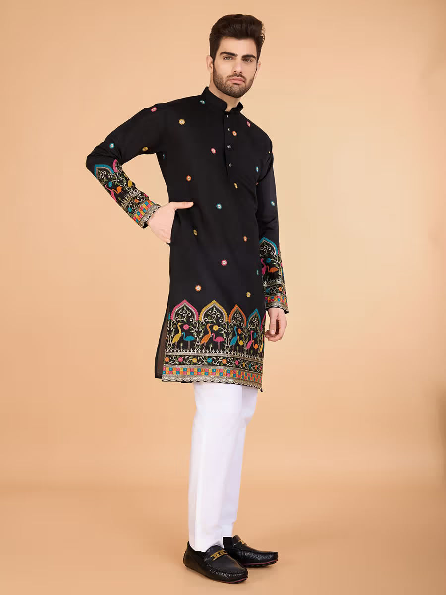 “Man wearing a black printed kurta with multicolor traditional patterns, paired with white pants, standing beside a wooden chair.”