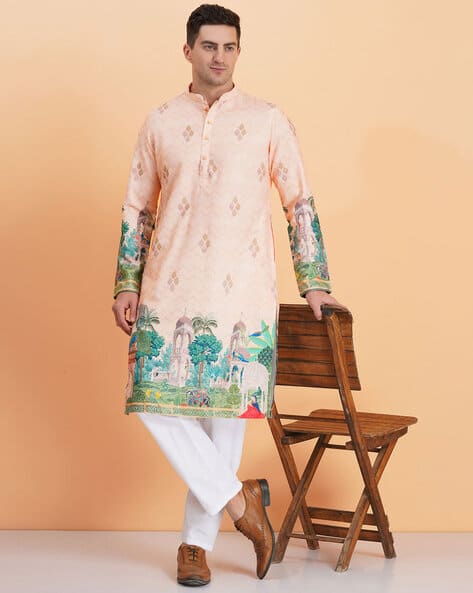 Man wearing a peach kurta with green sleeves and white pants, standing next to a wooden chair on a beige background.