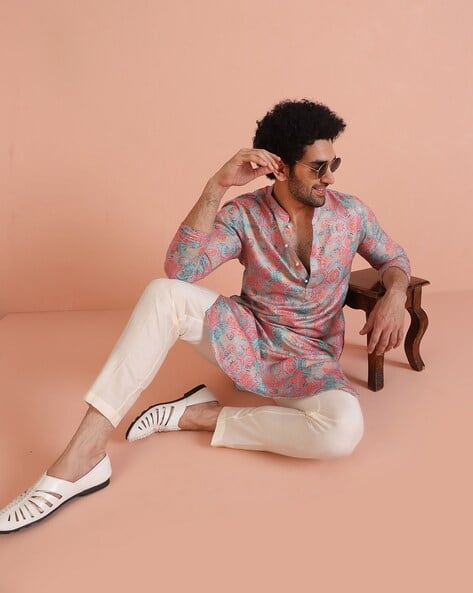 Man wearing a patterned shirt and white pants sitting on a stool against a peach background