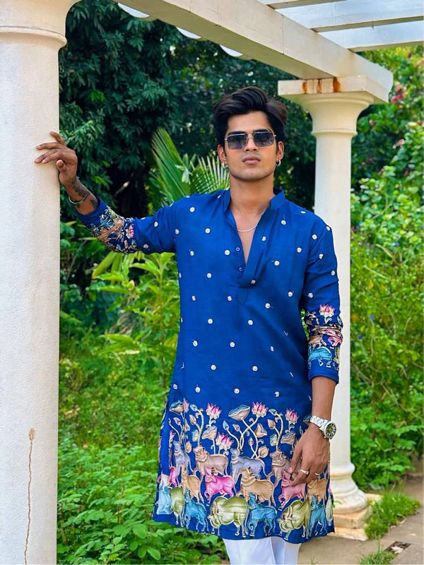 Man wearing a blue embroidered kurta with sunglasses, standing outdoors with greenery in the background